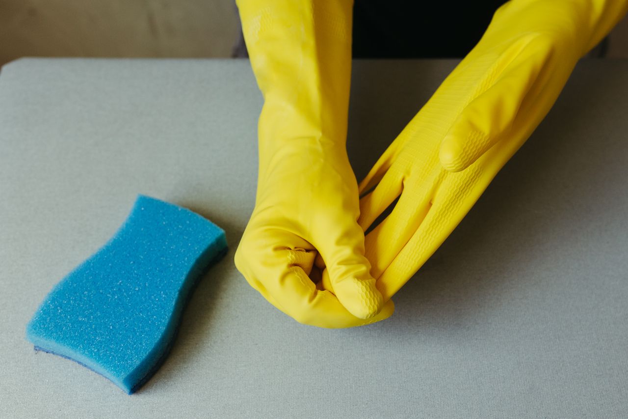 team-03 Close-up of hands wearing yellow gloves next to blue sponge on gray surface.